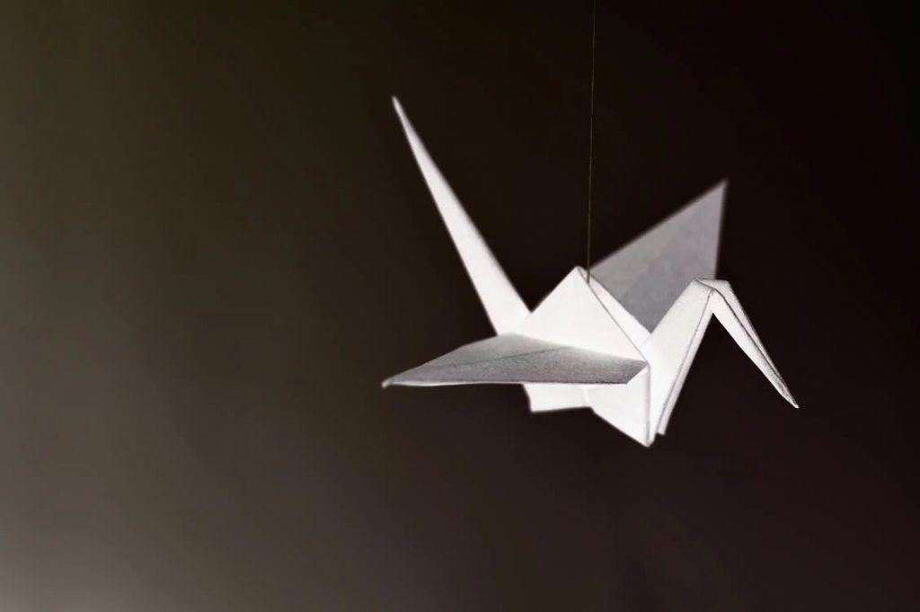 Żuraw origami złożony z białego papieru, skierowany w prawą stronę. Czarne tło.